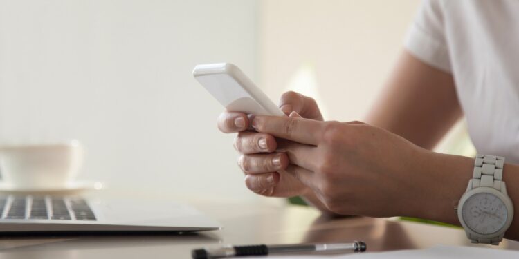 Close up image of modern cellphone in womans hand at desk with laptop. Office worker boring at workplace and playing games, writing in social networks. Businesswoman looking important contact on phone