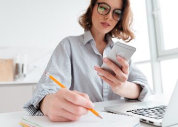 Cropped photo of serious young woman in glasses holding mobile phone and taking notes while sitting in light apartment, selective focus on hand