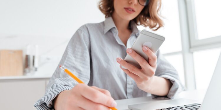 Cropped photo of serious young woman in glasses holding mobile phone and taking notes while sitting in light apartment, selective focus on hand