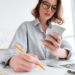 Cropped photo of serious young woman in glasses holding mobile phone and taking notes while sitting in light apartment, selective focus on hand