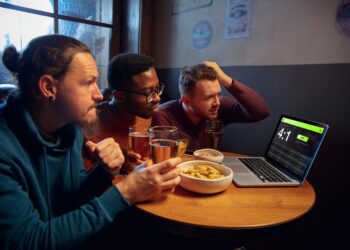 Device screen with application for betting and score. Device with match results on screen, excited fans on background during match. Gambling, betting, sport, finance, modern technologies concept.