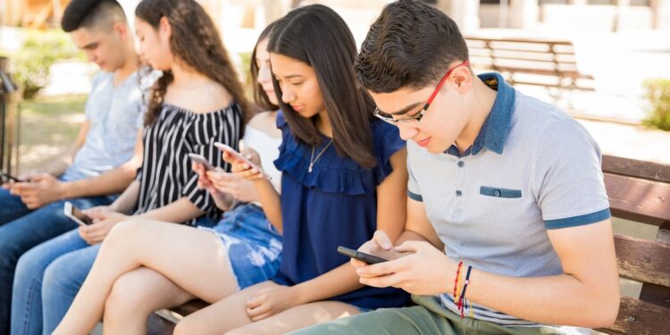 Young boy with friends using smart phones sitting on bench outdoors