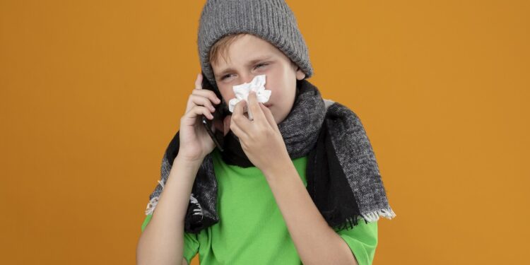 ill little boy wearing green t-shirt in warm scarf and hat feeling unwell talking on mobile phone wiping nose with paper napkin suffering from cold standing over orange background