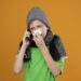 ill little boy wearing green t-shirt in warm scarf and hat feeling unwell talking on mobile phone wiping nose with paper napkin suffering from cold standing over orange background