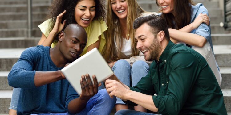 Multi-ethnic group of young people looking at a tablet computer outdoors in urban background. Group of men and woman sitting together on steps.