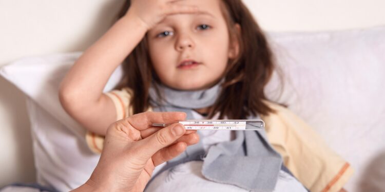 Mother holding thermometer in hands, sick little girl lying in bed and keeping her palm on forehead, looks sad and upset, suffering from high temperature, needs treatment and examining by doctor.