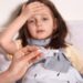 Mother holding thermometer in hands, sick little girl lying in bed and keeping her palm on forehead, looks sad and upset, suffering from high temperature, needs treatment and examining by doctor.