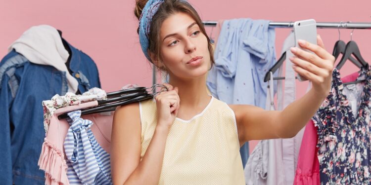 Pretty young female making selfie while standing near racks with clothes, being glad to spend her free time in shopping centre. Adorable lady using modern mobile phone while doing shopping alone.