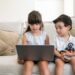 Focused pensive little brother and sister sitting on sofa at home, using laptop for video call or watching video or movie. Front view. Kids leisure time or internet concept