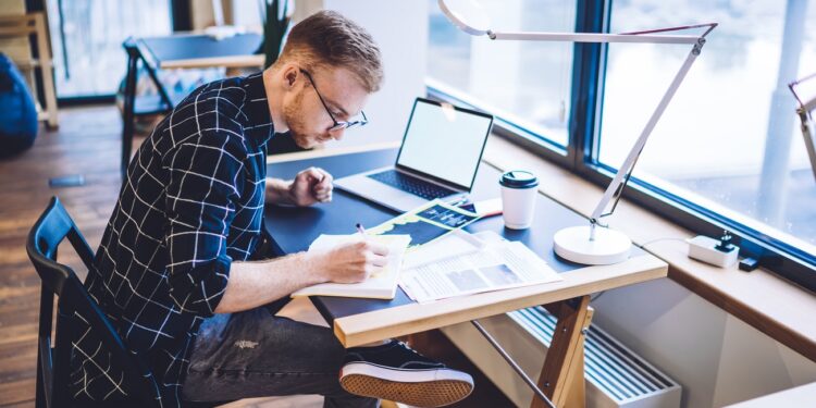 Caucaisan male student writing useful information notes in knowledge textbook sitting at coworking desktop with modern blank laptop with copy space area for internat website, concept of education