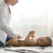 Beautiful young mother in white shirt and jeans putting diaper on cute newborn baby preparing for family dinner with grandparents. Spending special time with family