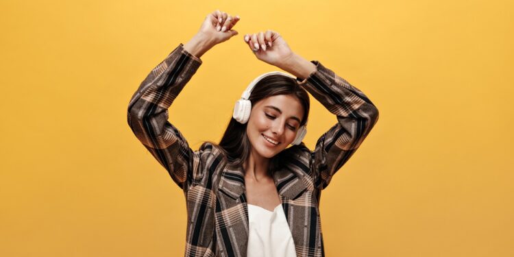 Joyful woman in white top, stylish brown jacket smiles and dances on yellow background. Attractive girl listening to music in headphones on isolated.