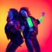 Portrait of young emotive people, man and woman wearing leather cloth, dancing isolated over gradient background in neon