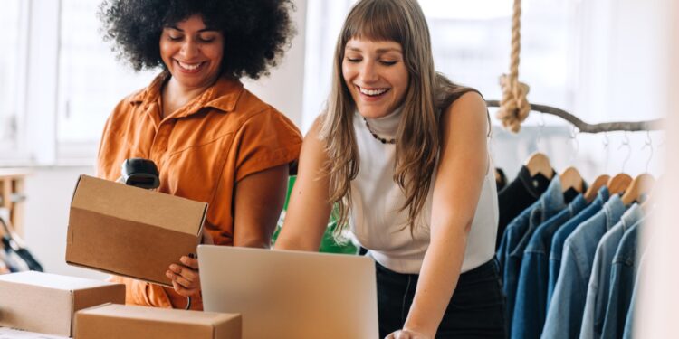 Happy businesswomen using a laptop while preparing delivery parcels in a thrift store. Two small business owners making plans for a shipment. Female entrepreneurs running an online clothing store.
