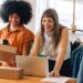 Happy businesswomen using a laptop while preparing delivery parcels in a thrift store. Two small business owners making plans for a shipment. Female entrepreneurs running an online clothing store.