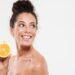 Close up portrait of a beautiful happy woman with soft healthy skin holding sliced orange and laughing isolated over white background