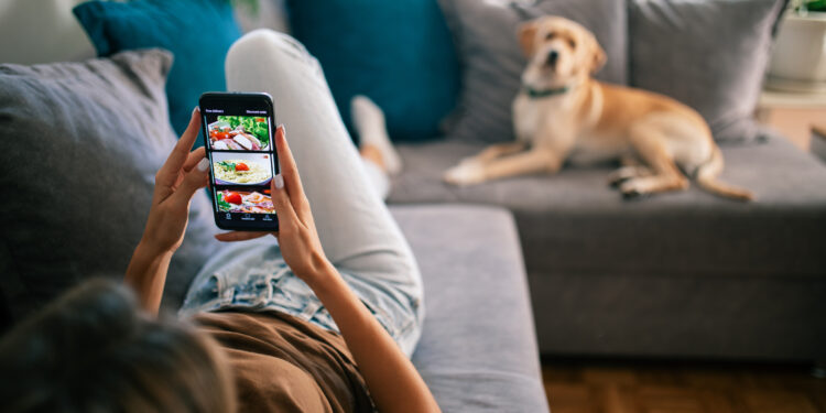 Young woman resting in her apartment on the sofa together with her Labrador retriever dog and ordering food for lunch online. She is holding a mobile phone and choosing meal to buy from the menu on the website. Photos used for app interface is my own photos, also available in my portfolio with photo IDs: 182682315, 182675346, 182702479