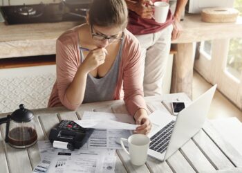 Couple facing debt problems, not able to pay out their mortgage. Thoughtful woman looking frustrated, holding pen while managing family budget, making calculations using calculator and notebook pc