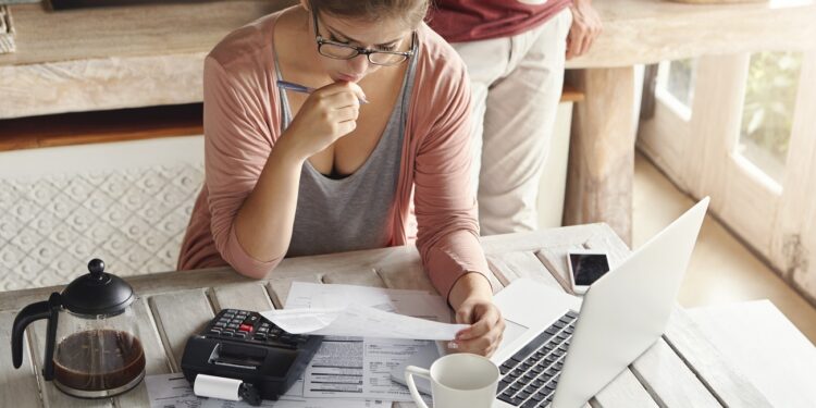 Couple facing debt problems, not able to pay out their mortgage. Thoughtful woman looking frustrated, holding pen while managing family budget, making calculations using calculator and notebook pc