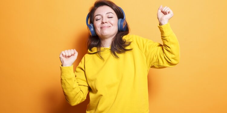 Excited lady dancing and listening to music on headset after winning a competition. Casual woman doing dance moves while using headphones to enjoy songs over isolated orange background.