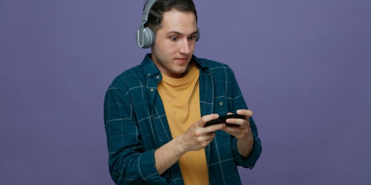 impressed young male student wearing headphones playing game on mobile phone isolated on purple background