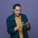 impressed young male student wearing headphones playing game on mobile phone isolated on purple background
