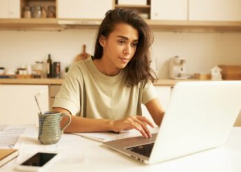 Indoor image of beautiful concentrated young female translator sitting in kitchen with mug, papers and portable computer translating technical document using free internet connection at home