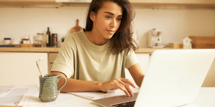 Indoor image of beautiful concentrated young female translator sitting in kitchen with mug, papers and portable computer translating technical document using free internet connection at home