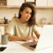 Indoor image of beautiful concentrated young female translator sitting in kitchen with mug, papers and portable computer translating technical document using free internet connection at home