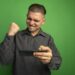 young handsome man in grey shirt using gadget clenching fist happy and excited rejoicing his success standing over green background