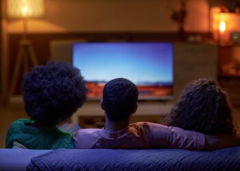 Back view of three people sitting on couch together and watching TV in dark room