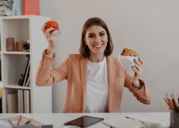 Lady in stylish light outfit shows off her lunch consisting of apple and burger in workplace