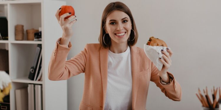Lady in stylish light outfit shows off her lunch consisting of apple and burger in workplace