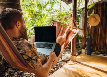 Man lounging in colorful hammock under palm trees using a laptop. Remote worker in relaxed outdoor environment.