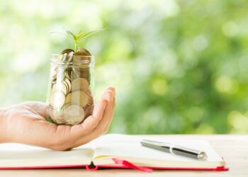 Woman hand holding plant growing from coins bottle in the on blurred green natural background