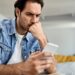 Young man feeling worried while reading text message on mobile phone at home.