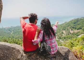 young happy hipster couple in love traveling around world, sightseeing, tropical romantic vacation in thailand, sitting on a rock, pointing finger, active tourism, summer