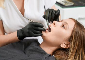 Female dentist in black sterile gloves examining patient teeth with dental explorer and mirror. Young woman lying in dental chair while having prophylactic examination. Concept of dentistry.