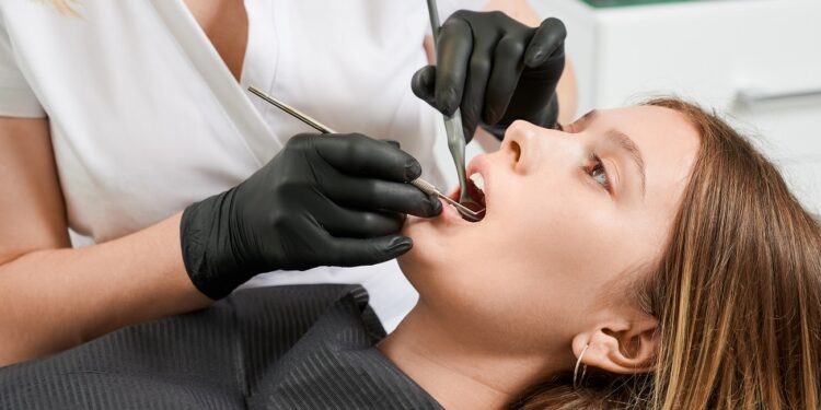 Female dentist in black sterile gloves examining patient teeth with dental explorer and mirror. Young woman lying in dental chair while having prophylactic examination. Concept of dentistry.