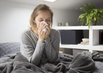 Sick young Caucasian woman covered with grey blanket sitting on bed with closed eyes, blowing nose with napkin. Illness, pain concept