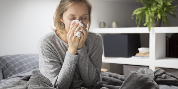 Sick young Caucasian woman covered with grey blanket sitting on bed with closed eyes, blowing nose with napkin. Illness, pain concept
