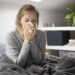 Sick young Caucasian woman covered with grey blanket sitting on bed with closed eyes, blowing nose with napkin. Illness, pain concept