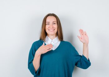 young woman keeping hand on chest, showing palm in sweater over white shirt and looking merry. front view.