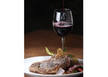 A vertical closeup shot of a freshly grilled T-bone steak and a glass of wine in the background