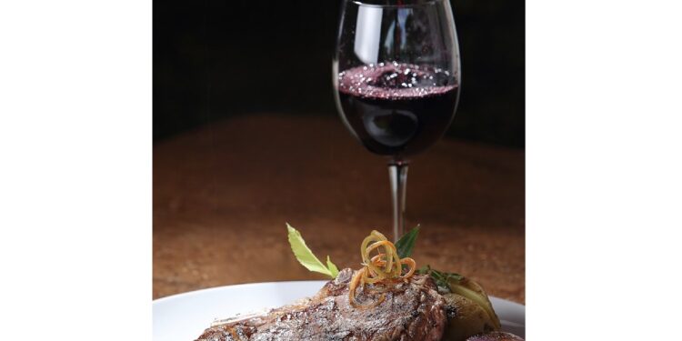 A vertical closeup shot of a freshly grilled T-bone steak and a glass of wine in the background