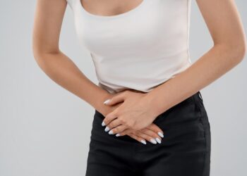 Clolse up of bending incognito caucasian woman in casual suit posing over gray isolated background and touching lower abdomen because of ache. Front crop of brunette suffering from pain in stomach.