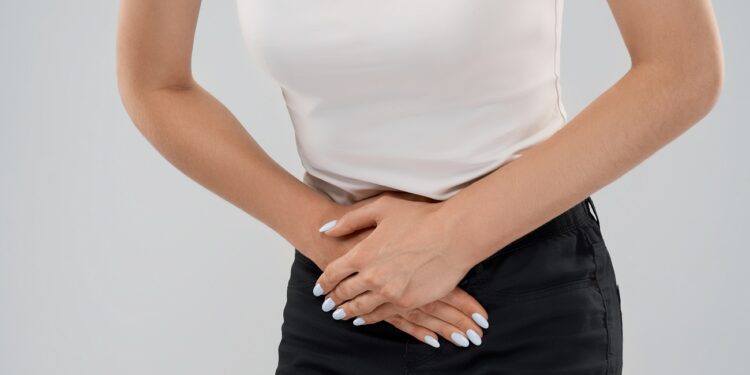 Clolse up of bending incognito caucasian woman in casual suit posing over gray isolated background and touching lower abdomen because of ache. Front crop of brunette suffering from pain in stomach.