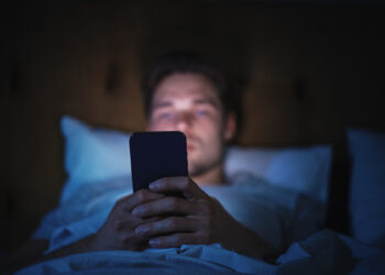Caucasian Man Uses Smartphone in Bed at Home at Night. Handsome Guy Browsing Social Media, Reading News, Doing Online Shopping, Chatting with Friends Late at Night. Focus on Hand Holding Mobile Phone