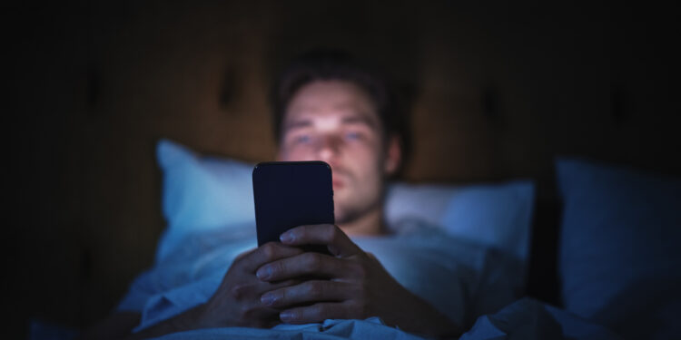 Caucasian Man Uses Smartphone in Bed at Home at Night. Handsome Guy Browsing Social Media, Reading News, Doing Online Shopping, Chatting with Friends Late at Night. Focus on Hand Holding Mobile Phone