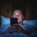 Caucasian Man Uses Smartphone in Bed at Home at Night. Handsome Guy Browsing Social Media, Reading News, Doing Online Shopping, Chatting with Friends Late at Night. Focus on Hand Holding Mobile Phone